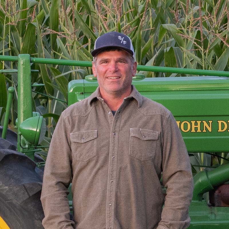 Man standing in front of a green tractor with a corn field in the background