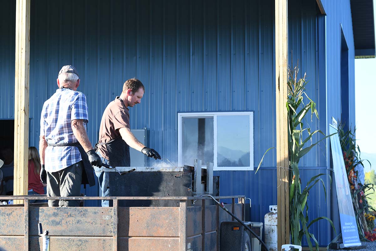 Two men working with a large grill outdoors against a blue barn.
