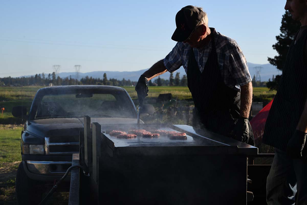 Man grilling meat on a portable grill outdoors with a truck in the background.