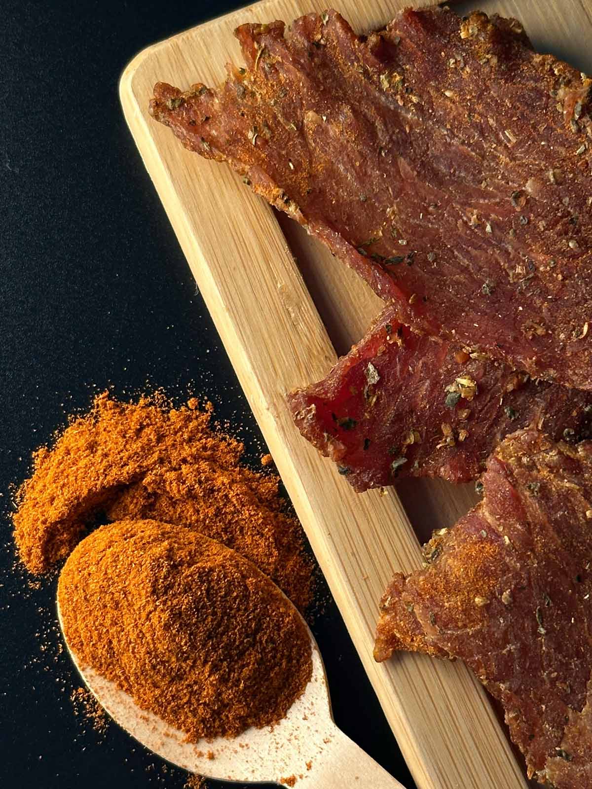 Dried meat slices on a wooden board with spices on a dark background