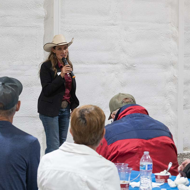 Woman in a cowboy hat speaking into a microphone to an audience against a white textured wall.