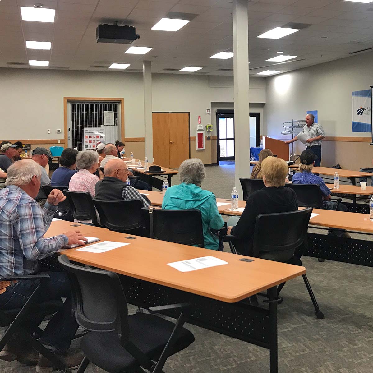 Group of farmers sitting at tables in a room with a speaker.